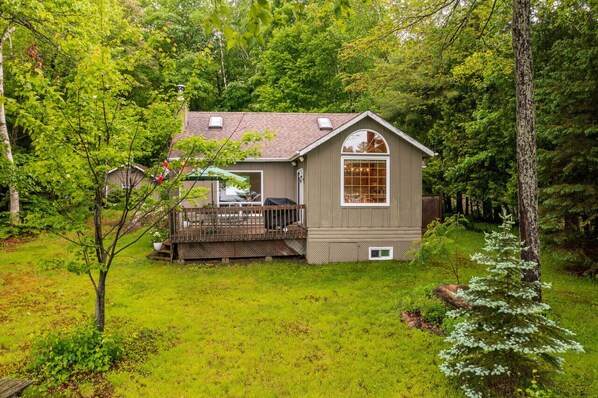 A small, single-story house with a deck, surrounded by lush greenery and trees.