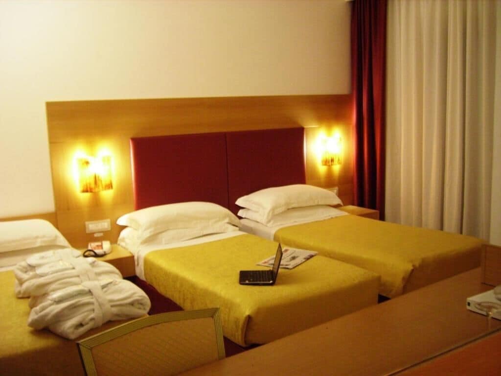 Hotellihuoneessa on kaksi erillistä sänkyä, joissa on kullankeltainen peitto ja valkoiset tyynyt. Kummankin sängyn vieressä on yöpöytä, jossa on lamppu, ja toisella sängyllä on kannettava tietokone.