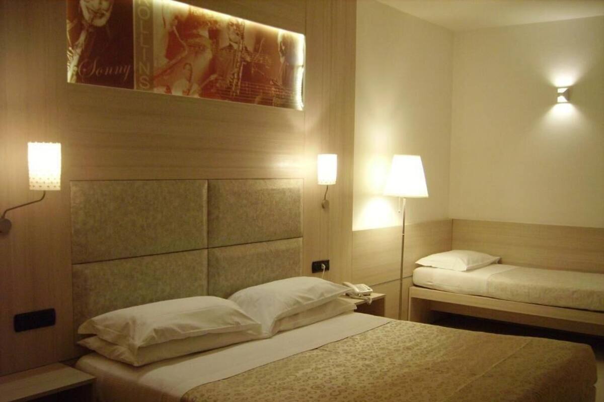 Habitación de hotel con una cama grande, otra más pequeña, una mesita de noche con lámpara y un teléfono en la pared.