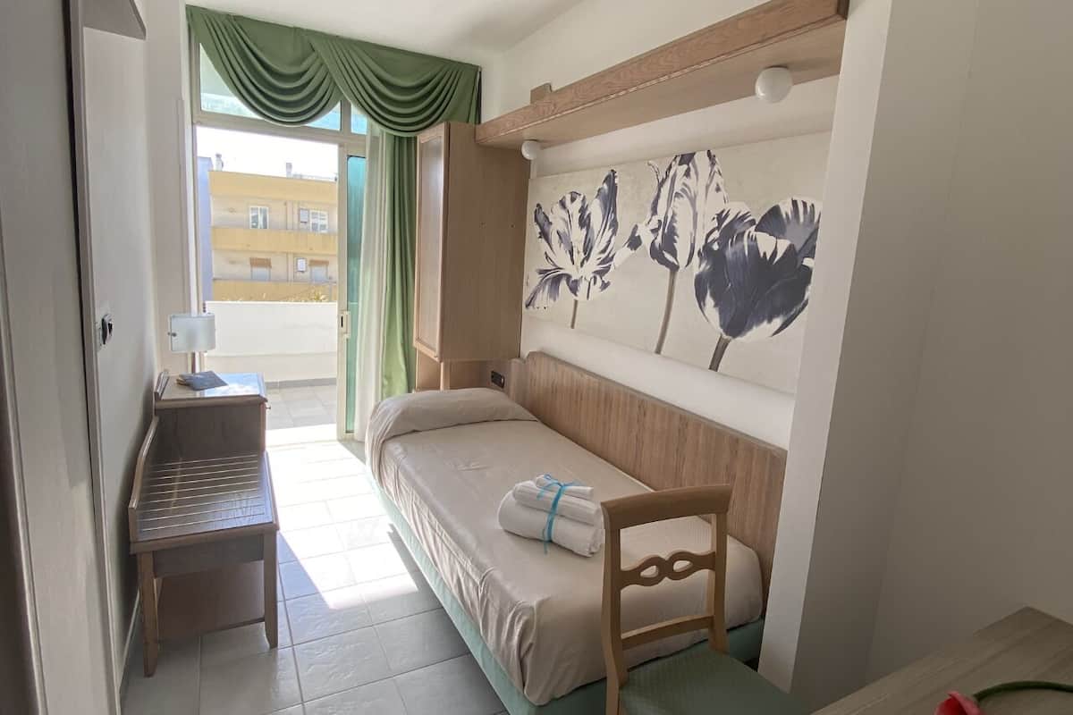 Una habitación pequeña y ordenada, con cama, mesita de noche de madera, estante en la pared y ventana con cortinas.