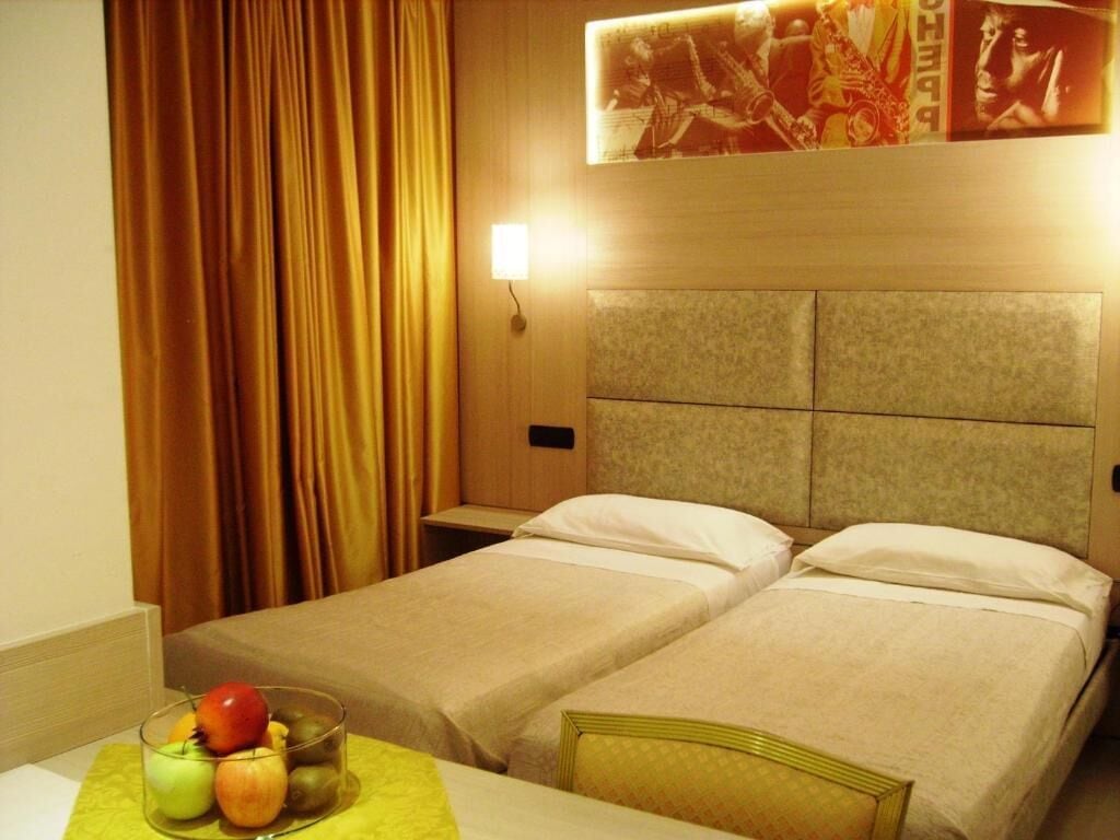 Una camera d'albergo con due letti, un comodino, una lampada e una ciotola di frutta sul tavolo.