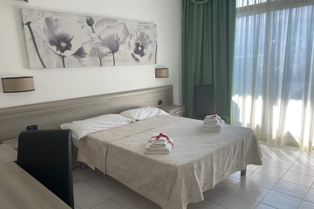 Habitación de hotel con cama, escritorio, silla, un cuadro en la pared y ventana con cortinas.
