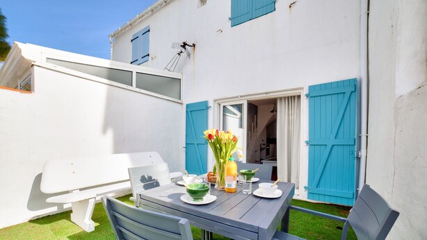 Un patio con una mesa para dos, una banca y una puerta con contraventanas azules.