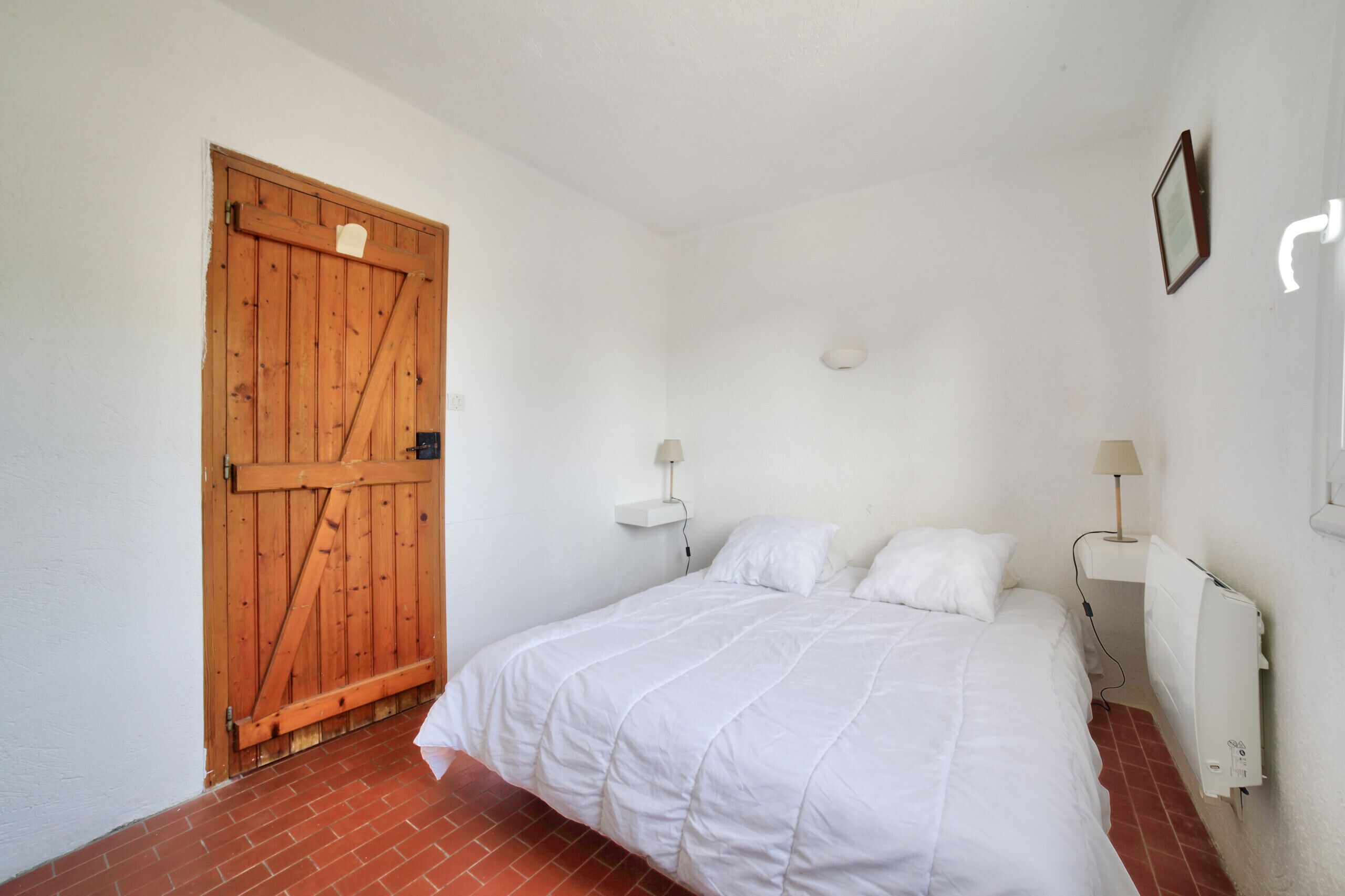 Un dormitorio con puerta de madera, una cama con ropa de cama blanca, dos mesitas de noche con lámparas y un radiador.
