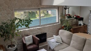 A spacious living room with a stone wall, a large window overlooking a body of water, a sofa, a coffee table, and a fireplace with a vase of flowers. - Villa Les Palmiers, 15 minutes from Monaco, Nice and the seaside. (Èze)