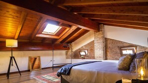 Slaapkamer in loftstijl met schuin dak, zichtbare houten balken, een haard van baksteen en een groot bed met uitzicht op de lucht via dakramen.