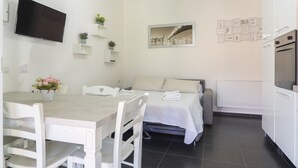 Ein modernes, helles Zimmer mit Esstisch, Stühlen, einem Bett, einem Fernseher und einer Küchenzeile.