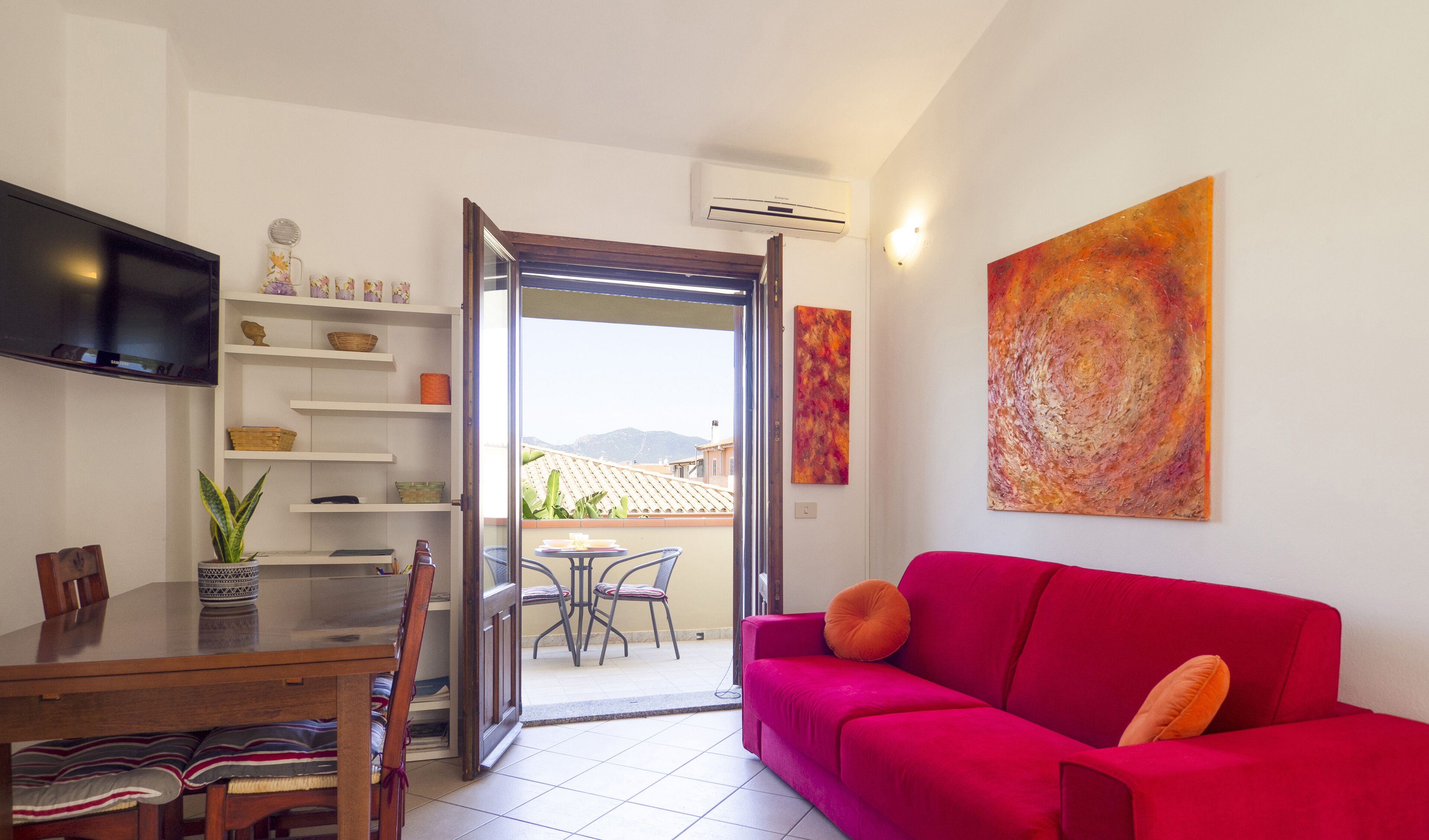 Un salon avec un canapé rouge, une table à manger avec des chaises, un téléviseur fixé au mur et un balcon offrant une vue sur les montagnes.