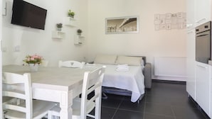 A modern, well-lit room with a dining table, chairs, a bed, a TV, and a kitchen area.