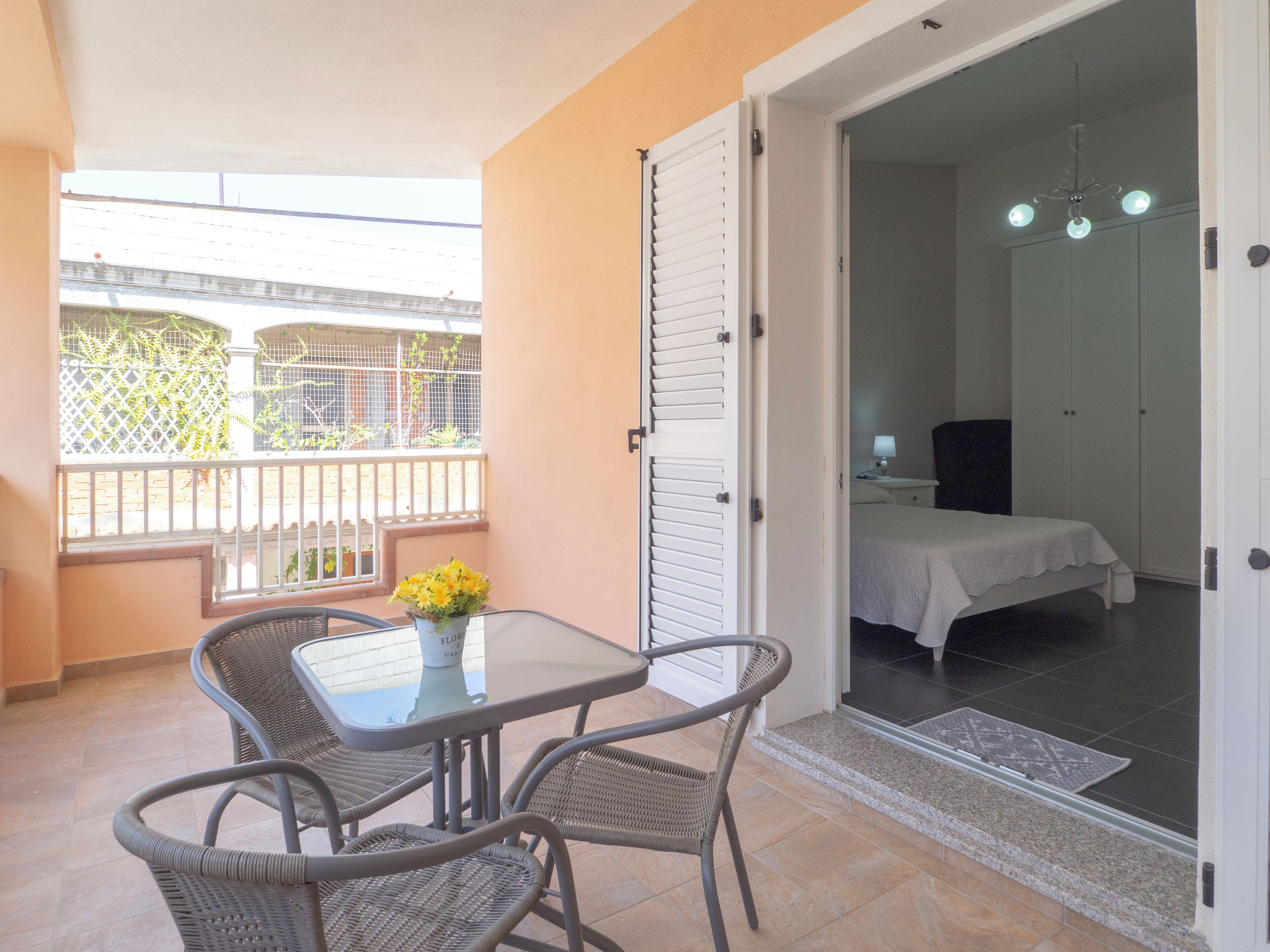 A balcony with wicker chairs and a table, a bed in the adjacent room, and a view of a building with a fence.