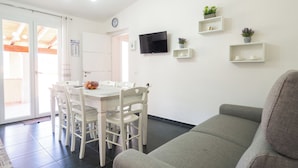 A modern living room with a grey sofa, a dining area with white chairs and a table, and a TV mounted on the wall.
