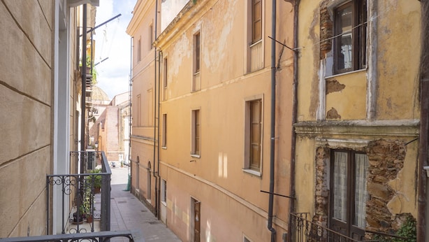 Une rue étroite, bordée de bâtiments des deux côtés, avec des balcons dotés de garde-corps et de plantes en pot.