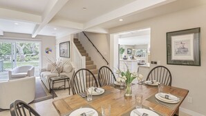 A dining area with a wooden table set for six, surrounded by chairs. A staircase is visible in the background leading to an upper level. - 3885 Pebble Beach Oasis - Indoor / Outdoor Living (Pebble Beach)