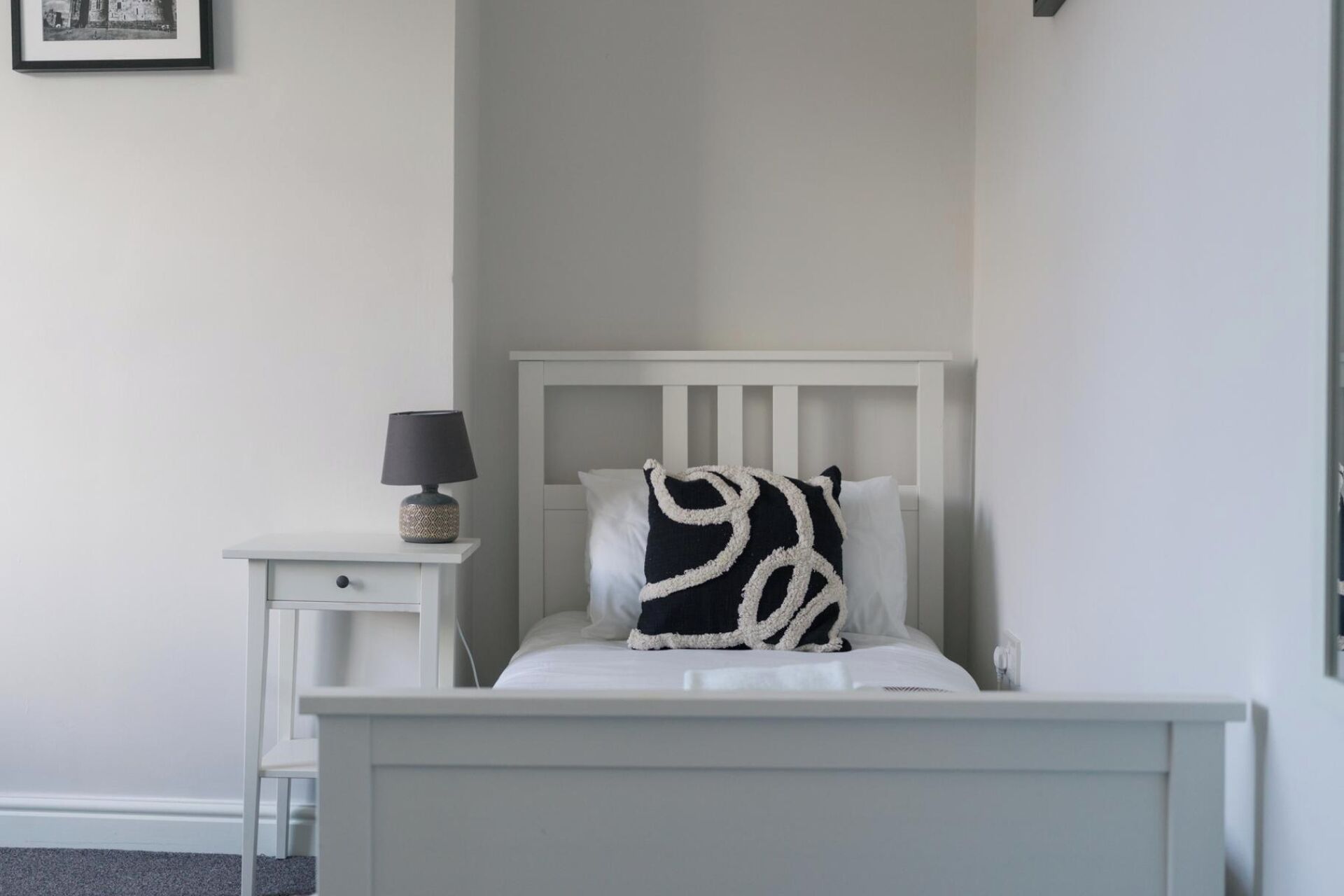 Un dormitorio con cama blanca, mesita de noche con lámpara y un cuadro en la pared.