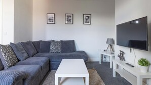 Uma sala de estar moderna com um sofá modular cinza, uma mesa de centro branca, uma TV de tela plana na parede e uma mesinha lateral com uma luminária e objetos decorativos.