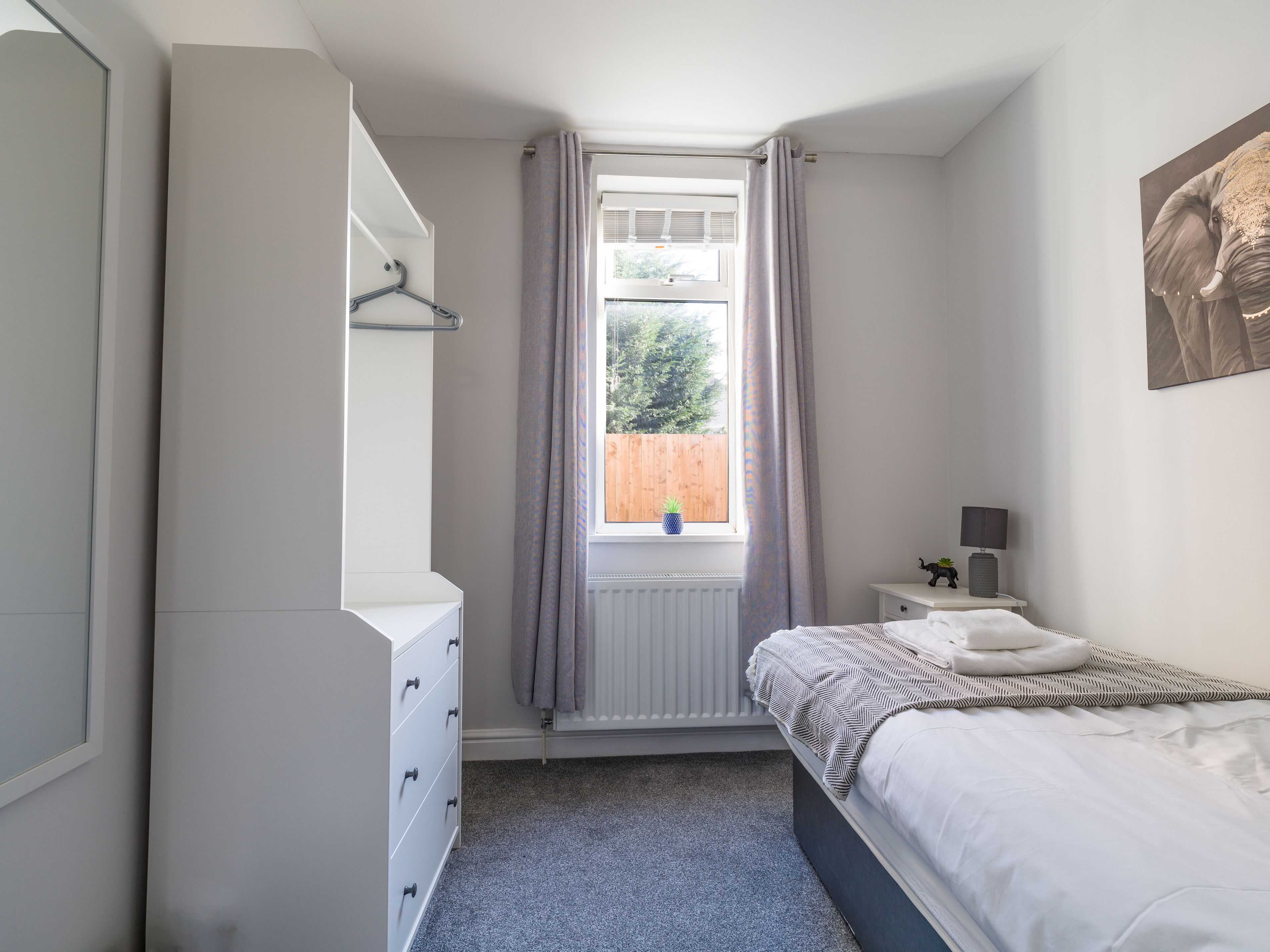 A bedroom with a bed, a dresser, a window with curtains, a radiator, and a small table with a lamp.