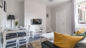 A modern, well-lit living room with a white sofa, a dining table set with plates and glasses, a TV mounted on the wall, and a window with curtains. - Lily Apartment 1- 2bed in Bedlington (Bedlington)
