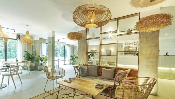 A modern living area with a sofa, chairs, a coffee table, and a dining table. There are wicker lamps, a large mirror, and a kitchen area visible in the background. - Brand new luxury studio condo,with private pool in Tuk  (Tulum)