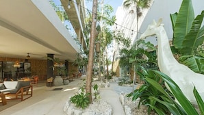 A modern indoor space with a central pathway, surrounded by seating areas and lush greenery, including a large giraffe sculpture. - Brand new luxury studio condo,with private pool in Tuk  (Tulum)
