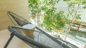 A balcony with a wicker chair and a small round table, overlooking a garden with trees and a building in the background. - Brand new luxury studio condo,with private pool in Tuk  (Tulum)
