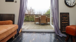 A room with a sliding glass door leading to an outdoor deck, a brown leather sofa, a wooden table with chairs, a clock, and a sign on the wall. - Hillcroft (St Austell)