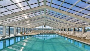 Piscina interna com tetos de vidro transparente, espreguiçadeiras e vista para a cidade.