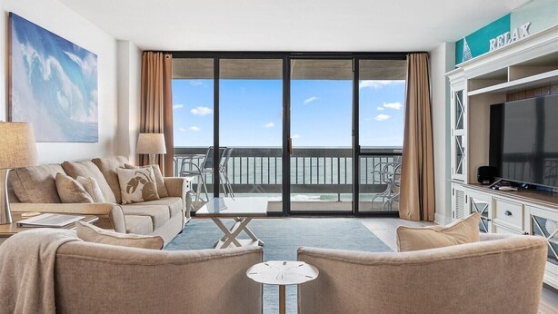 Sala de estar moderna com uma televisão grande, assentos confortáveis, uma varanda com vista para o oceano e um letreiro com a palavra 'RELAX'.