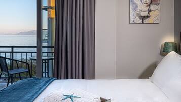 A hotel room with a bed, a view of the sea, a painting on the wall, and a balcony with a chair and table.