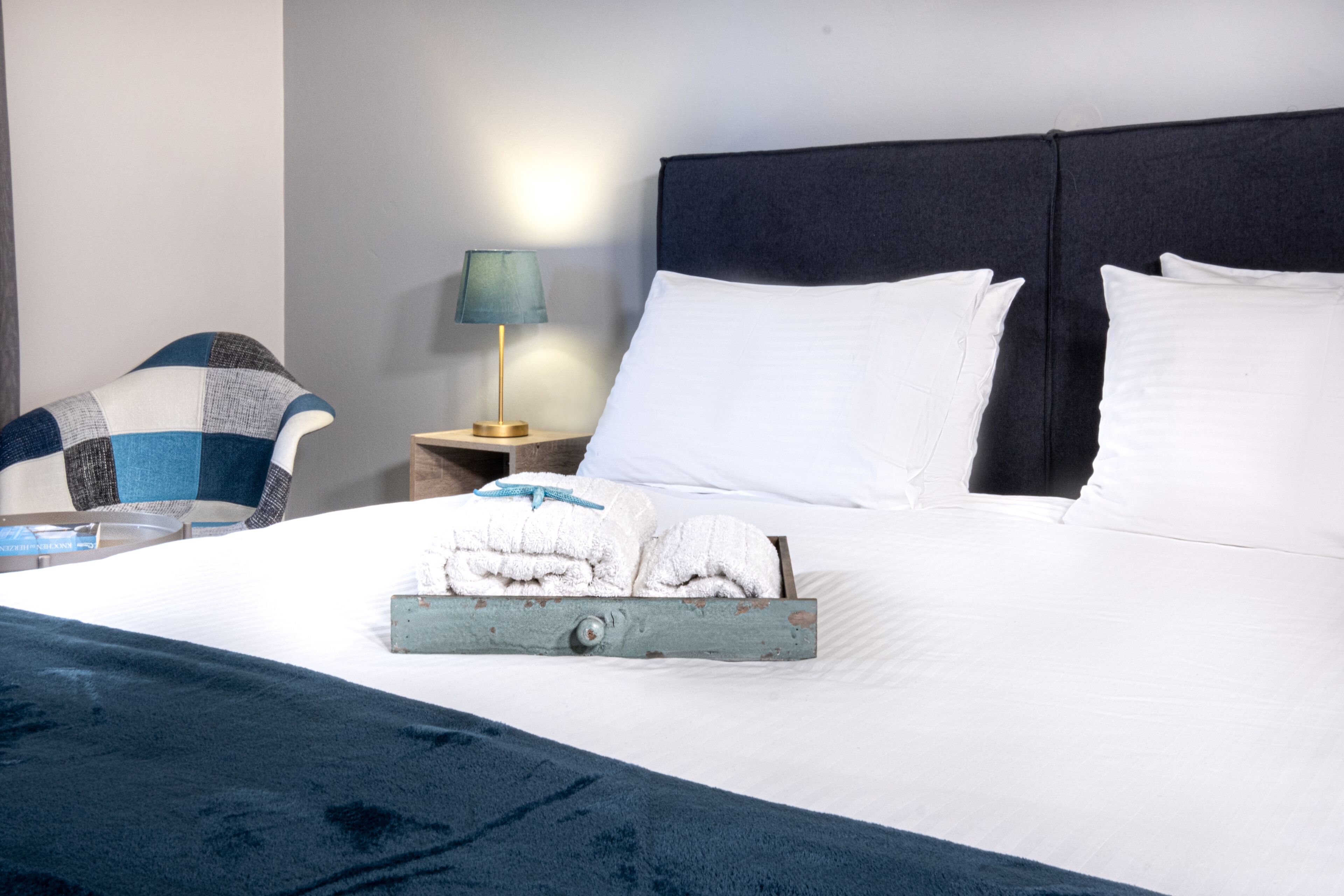 Un lit bien fait, avec du linge de lit blanc, une couverture bleue et un plateau contenant des serviettes pliées. On y trouve une table de chevet en bois avec une lampe, ainsi qu’une chaise à motifs bleus et blancs.