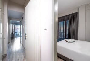 A hotel room with a bed, a window with curtains, a view of the outdoors, and a hallway leading to another room.