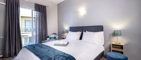 Habitación de hotel moderna con una cama grande, dos mesitas de noche, un taburete pequeño y vistas al balcón exterior.