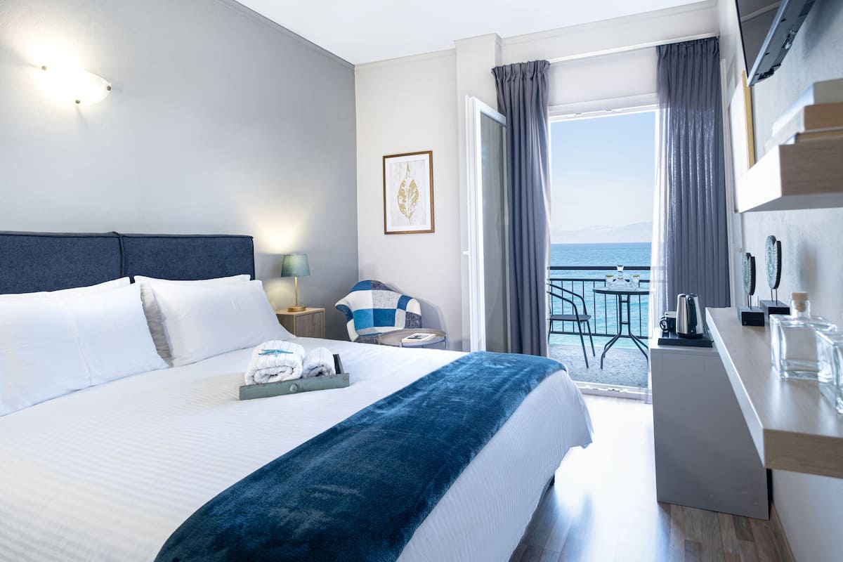 A hotel room with a bed, a desk, a chair, a table, a balcony with a view of the sea, and a window with curtains.