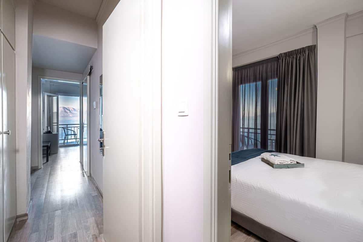 A hotel room with a bed, a window with curtains, a door, and a hallway leading to another room.