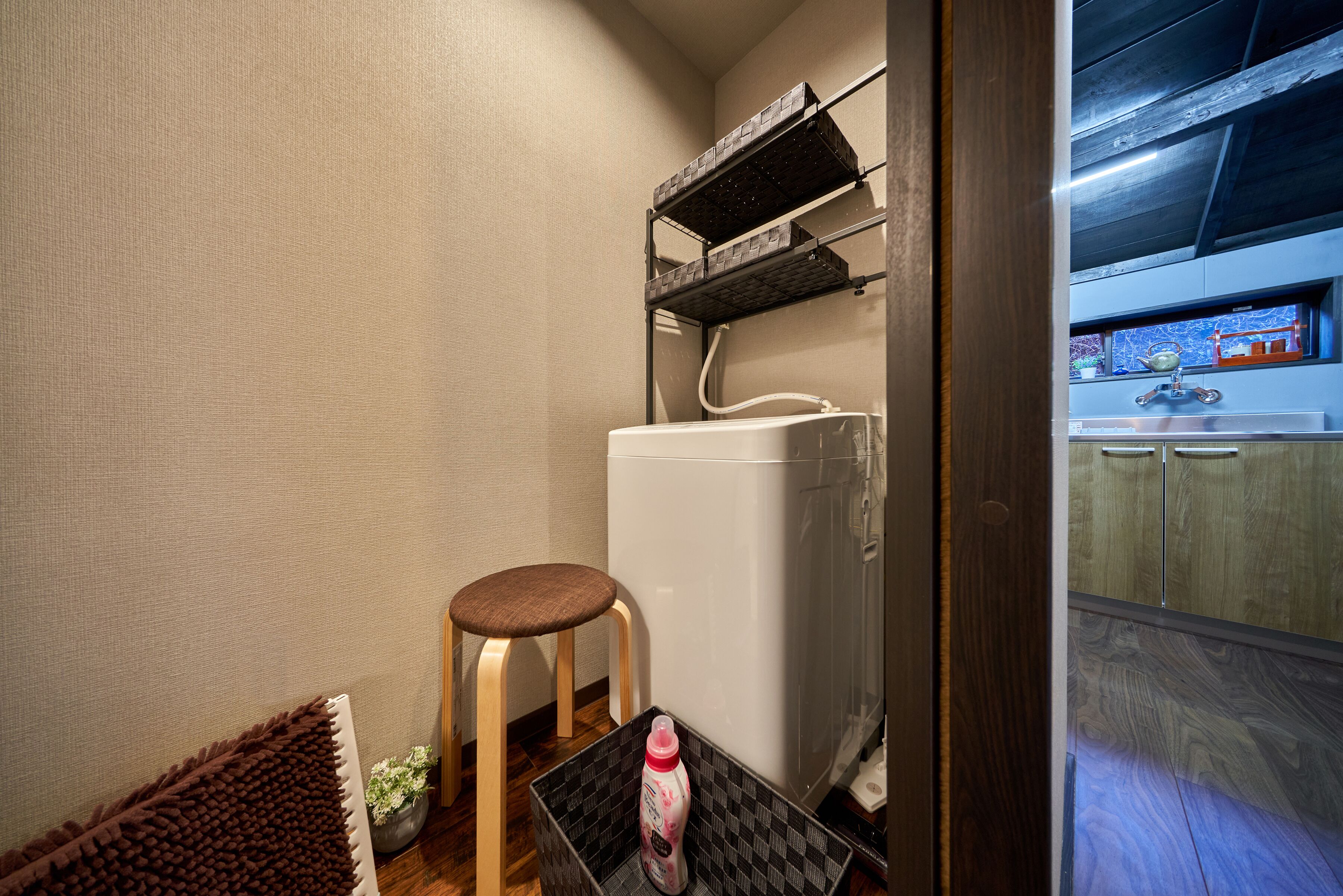 A compact bathroom with a white toilet, a wooden stool, a small table with a bottle, and a shelf with toiletries.