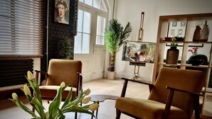 Ein Wohnzimmer mit zwei Sesseln, einem Couchtisch und einer Vase mit Blumen. An der Wand hängt ein Gemälde und es gibt ein Fenster mit Jalousien.