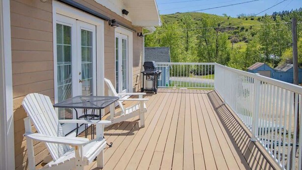A wooden deck with outdoor seating, a grill, and a view of a green hillside. - Panoramic Platt- Mountain and River Views- In Town (Red Lodge)
