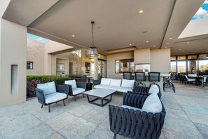 An outdoor patio with modern furniture, including sofas and chairs, a dining area, and a kitchen visible through the glass walls.