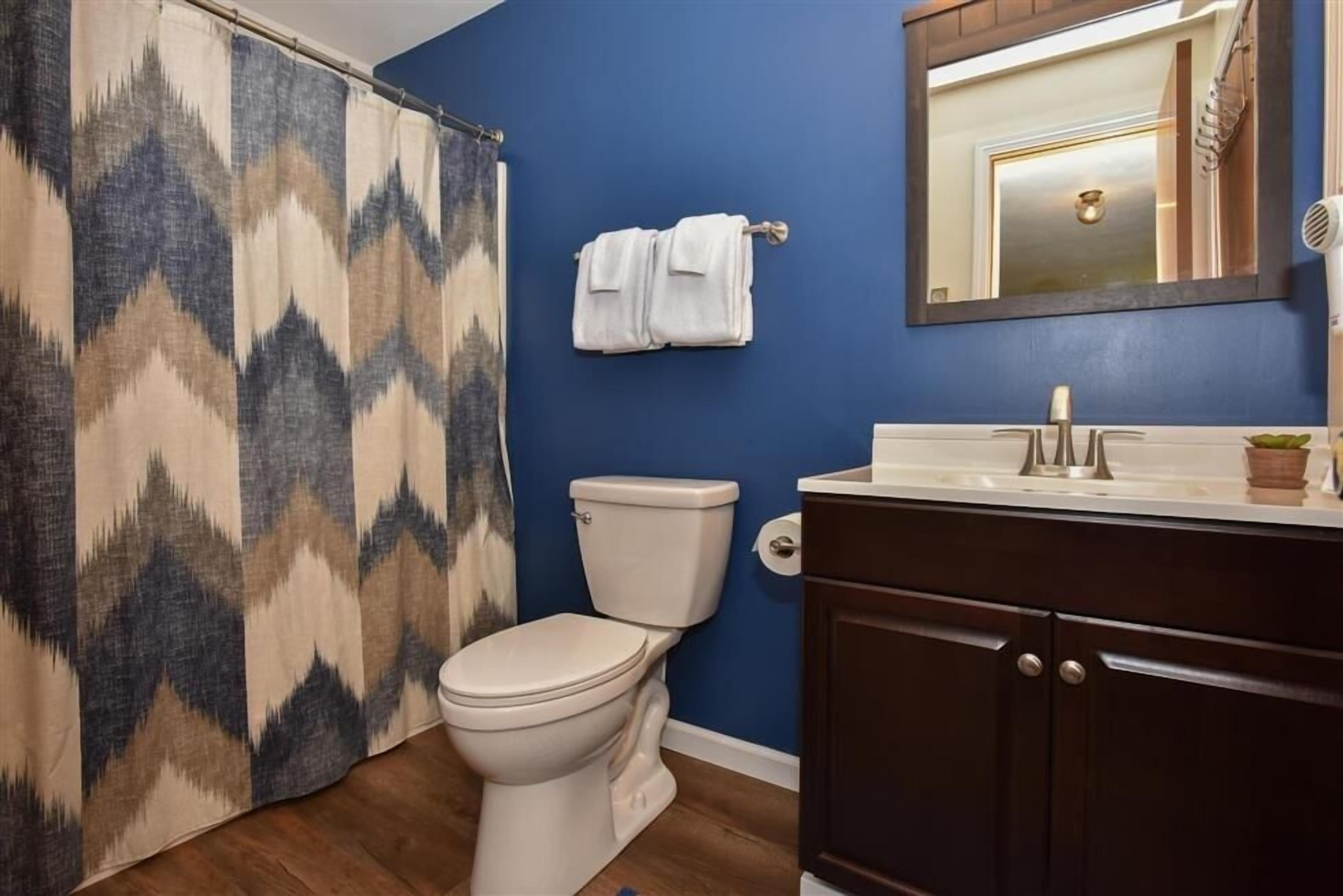 Ein Badezimmer mit einer weißen Toilette, einer blauen Wand, einem dunklen Holzschrank und einer Duschvorhang mit einem geometrischen Muster.
