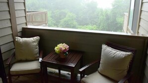 A balcony with two chairs and a small table, a potted plant, and a view of trees through the window.