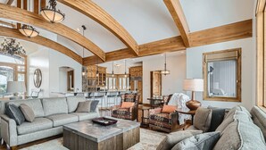 A spacious living room with a large sectional sofa, a coffee table, and patterned rugs. The room features exposed wooden beams, pendant lighting, and a view of the kitchen and dining area.