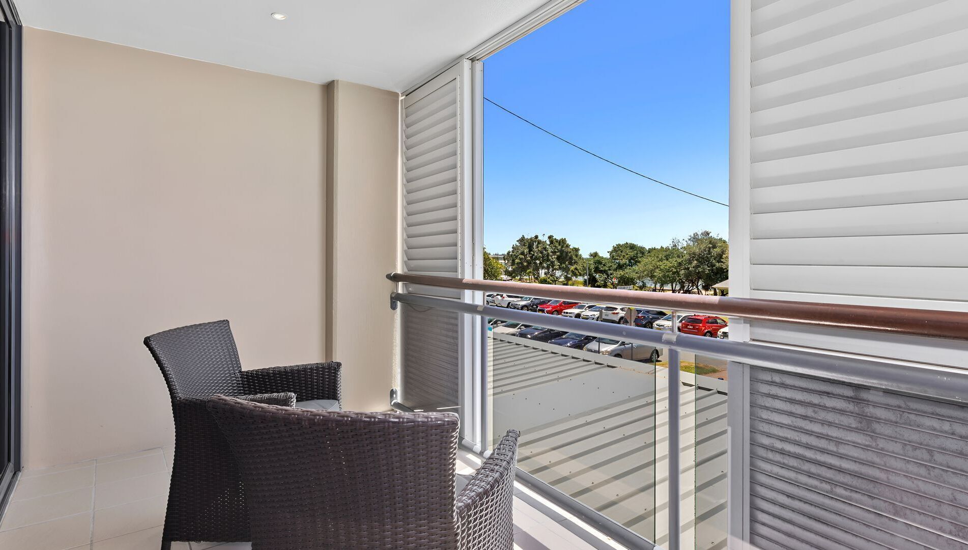 Un balcon avec des fauteuils en osier, une vue sur un parking et un ciel bleu dégagé.