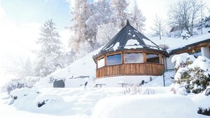 En træhytte med et kegleformet tag, omgivet af sneklædte træer og en klar himmel.