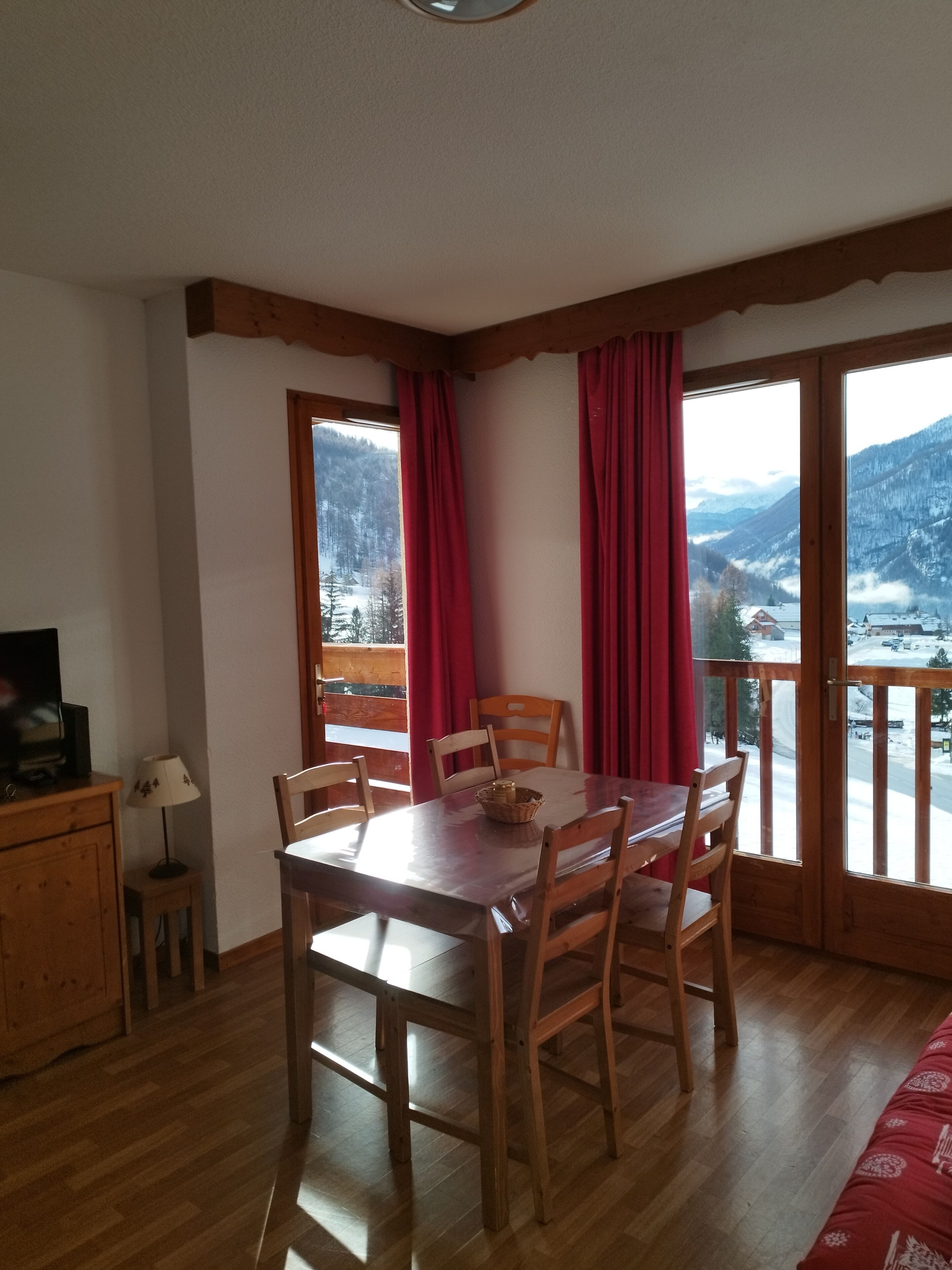 Una zona de dinar amb taula i cadires de fusta, un moble petit i una làmpada. Hi ha grans finestres amb cortines vermelles, que donen vista a un paisatge de muntanyes nevades.