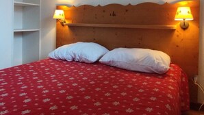 Una cama con una colcha floral roja y dos almohadas blancas, flanqueada por dos lámparas fijadas a la pared.