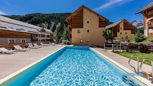 Una piscina con sillas de descanso, edificios de madera y montañas al fondo.