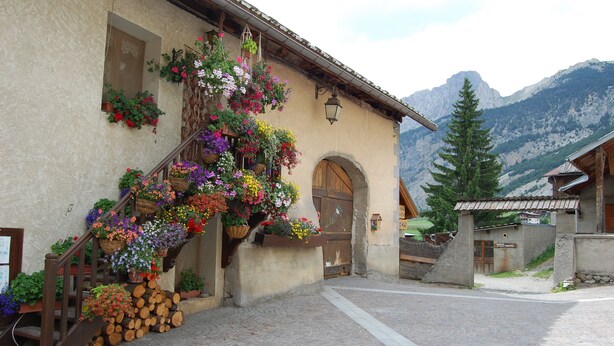 Un edificio con una escalera de madera que lleva a una puerta, rodeado de cestas de flores y leña.