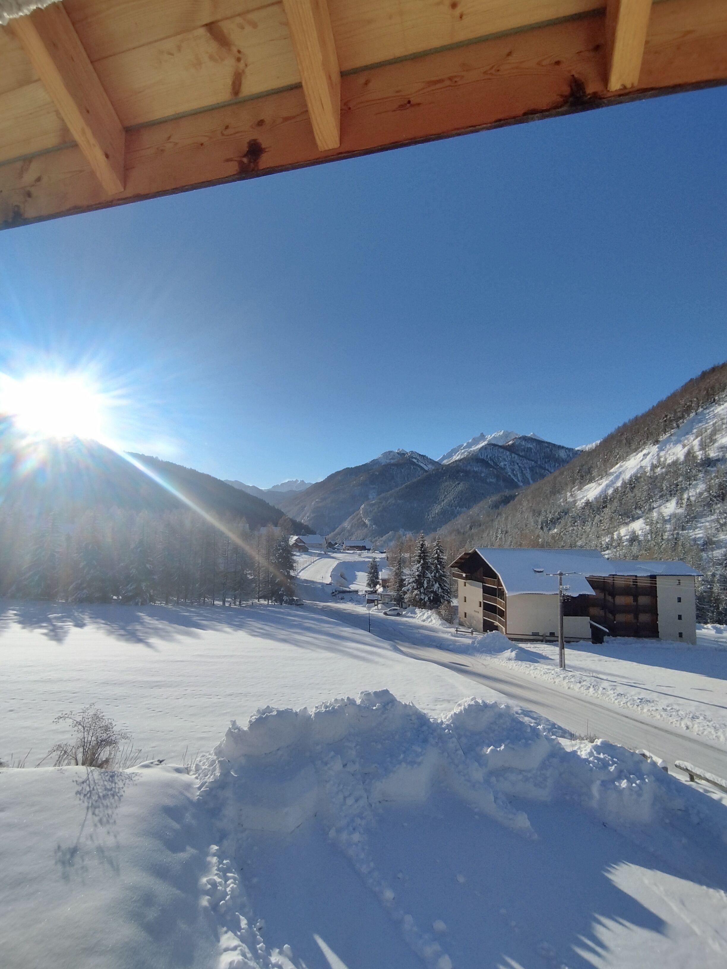 A snow-covered landscape with a clear sky, mountains in the background, and a building with a sloped roof.