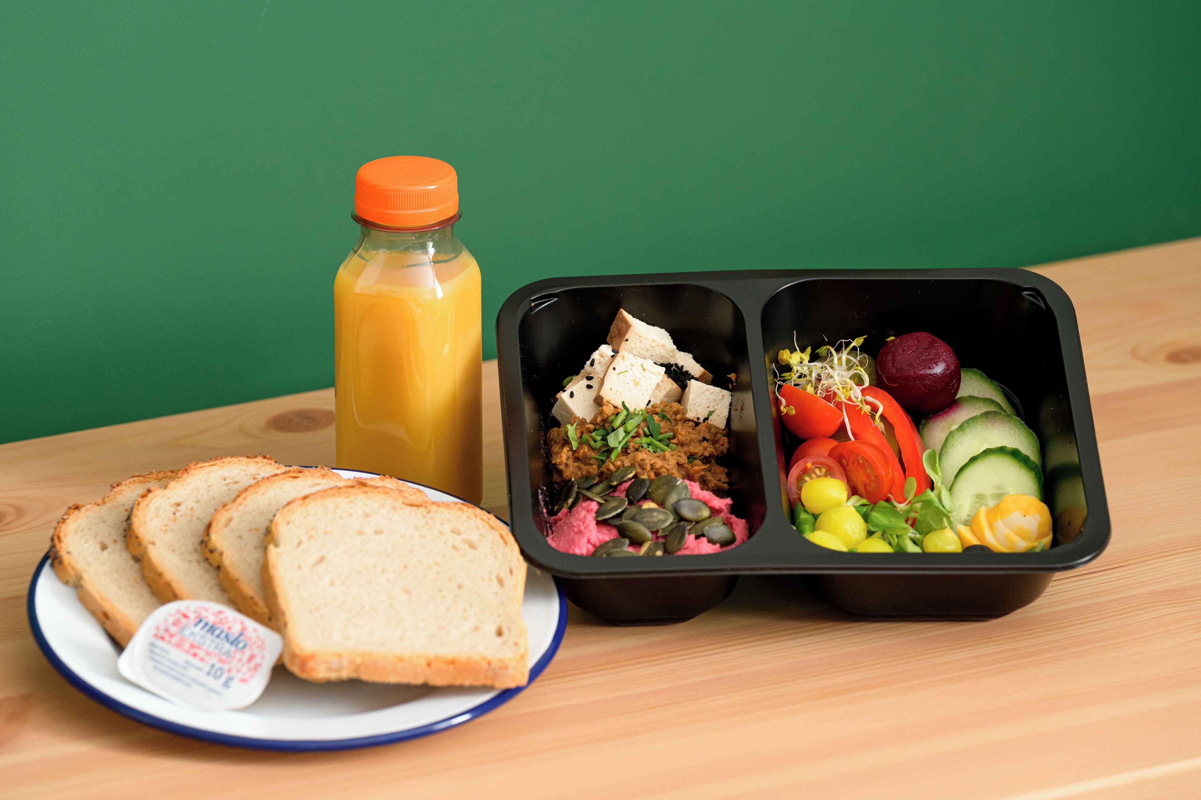 A meal with bread, a juice bottle, and a salad in a black container on a wooden table.