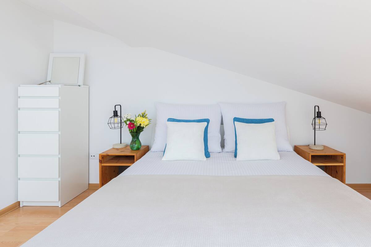 Une chambre à coucher avec un lit, des murs blancs, une table de chevet en bois, une commode, un petit meuble en bois et un vase avec des fleurs.
