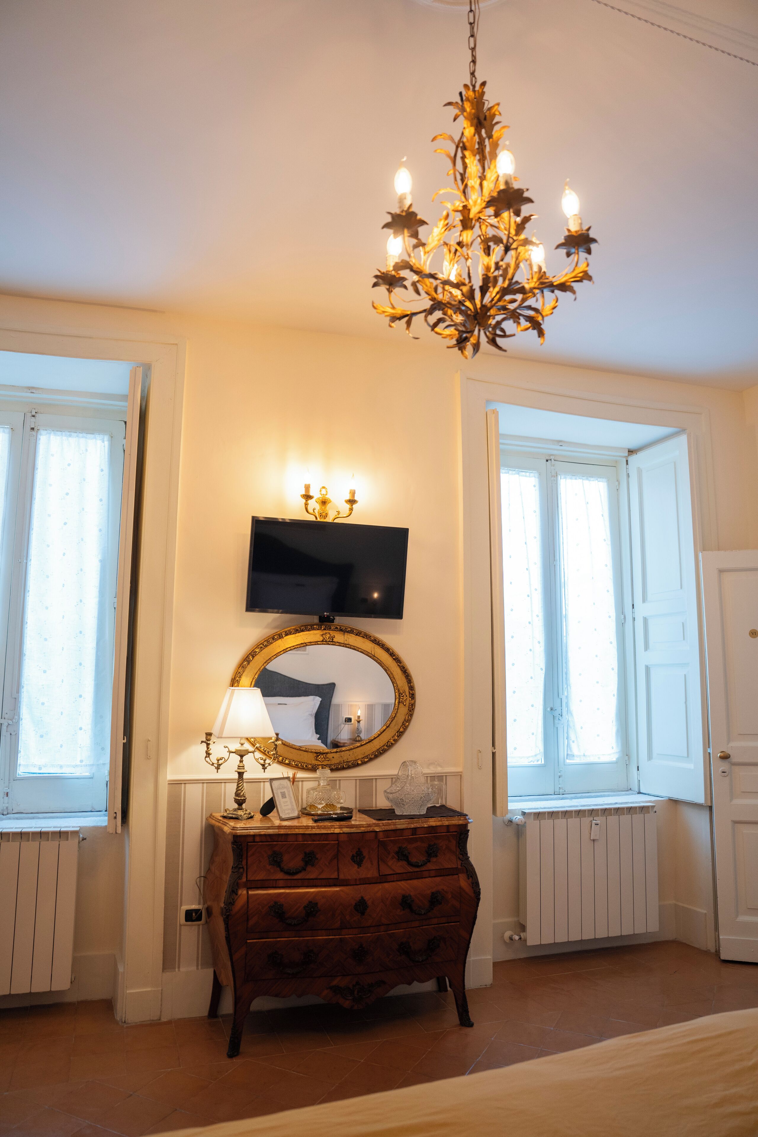 A room with a chandelier, a television mounted above a mirror, a dresser, and two windows with open shutters.